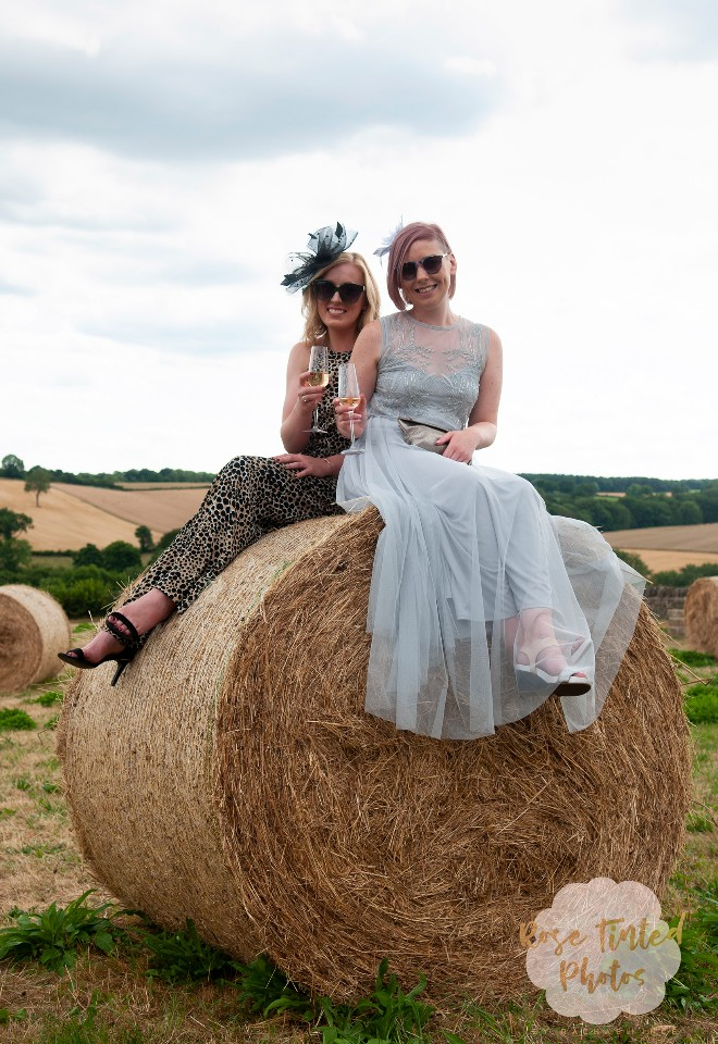 Two adventurous wedding guests sit atop a haybale in the countryside, champagne flutes in hand, sunglasses on and smiling, clearly pleased with their climbing efforts
