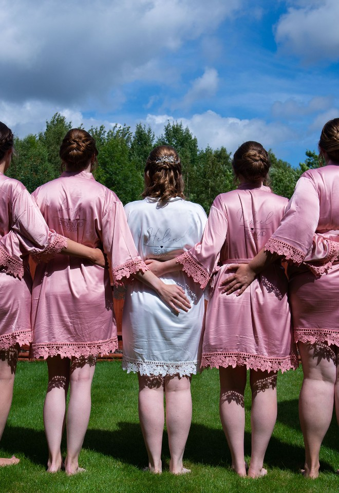 Bride and 4 bridesmaids stand in her garden with their backs to camera, arms around each other, showing off their bride and bridesmaid dressing gowns