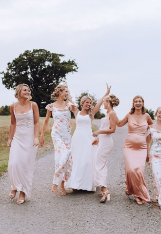 Bride and her bridesmaids wearing mismatched dresses in pink and white tones. They are walking down a driveway surrounded by countryside.