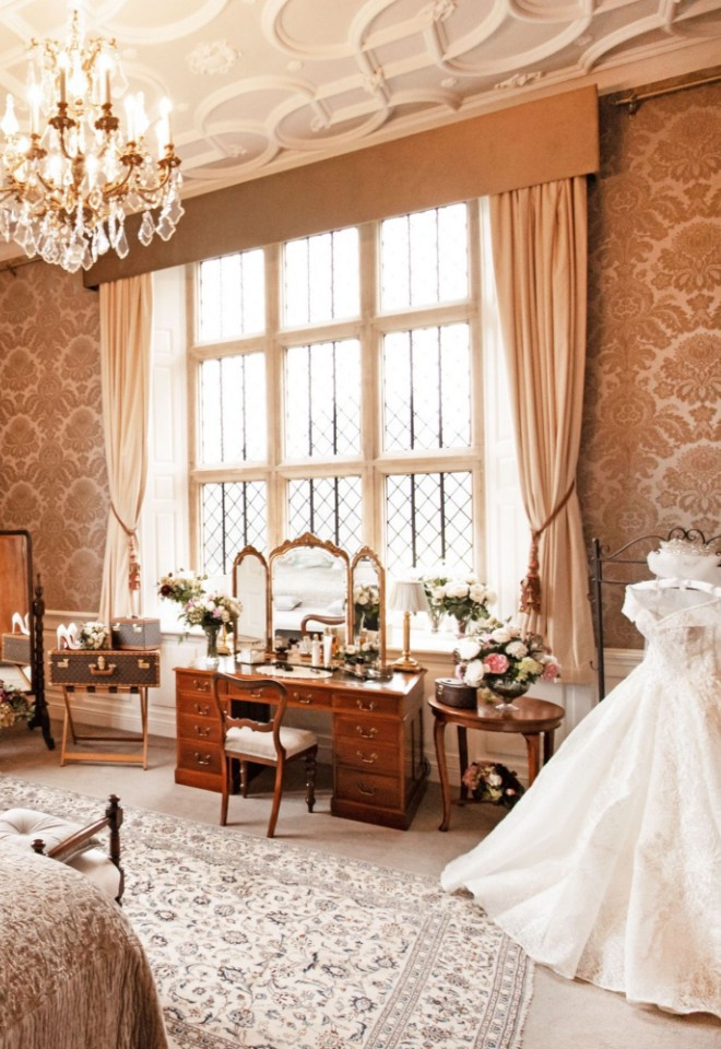 Bridal suite at North Mymms Park featuring large leaded windows, a crystal chandelier, a vintage vanity, and a white wedding dress on display.