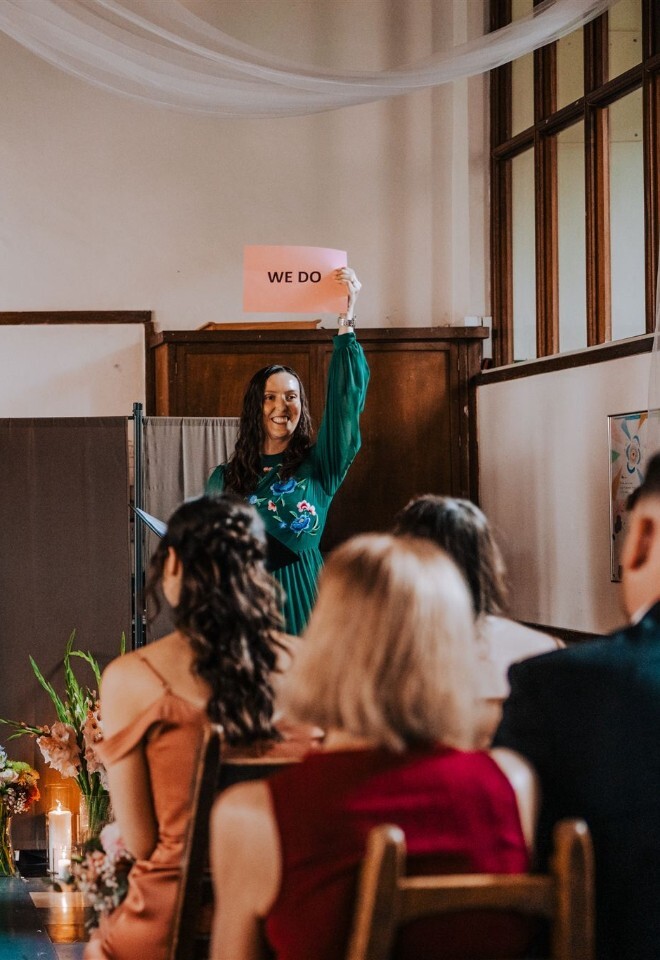 UK wedding celebrant holding up a card saying We do while leading guest group vows