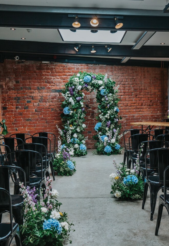 Blue and purple flower arch at the top of the aisle in front of exposed brick wall