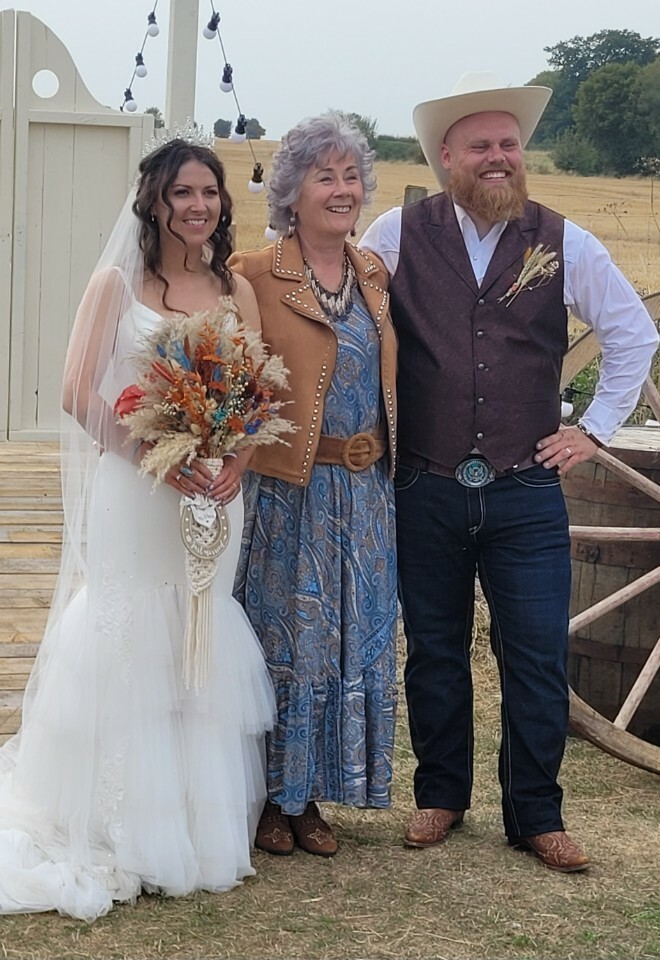 Essex wedding celebrant dressed in boho outfit in between the bride and groom after their county music festival wedding ceremony