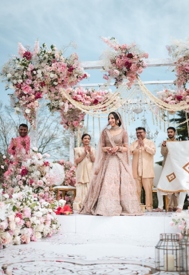 Bride in a detailed blush pink lehenga walking down the aisle towards an outdoor mandap adorned with lush pink and white flowers during a wedding ceremony at North Mymms Park.