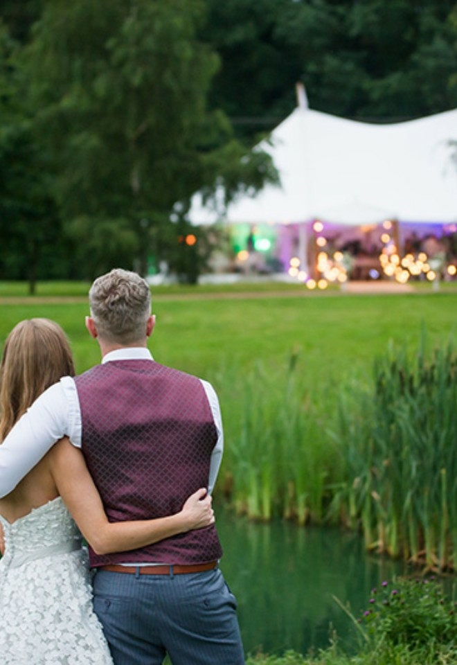 A newlywed couple embraces by a pond, overlooking a softly lit white marquee tent in the distance.
