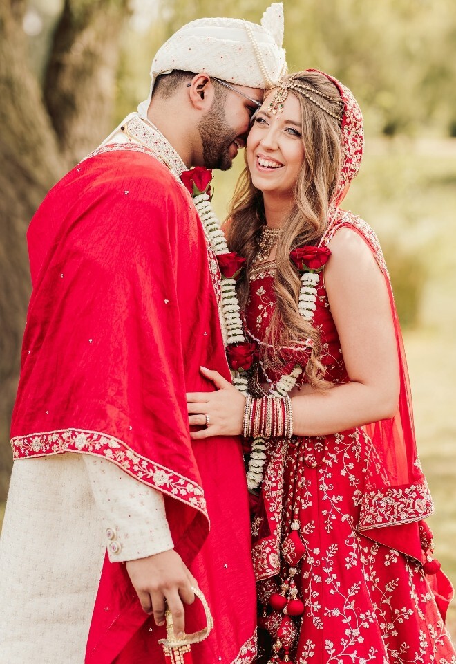 A couple in traditional Indian wedding attire, with the groom wearing a cream sherwani and red shawl and the bride in a red embroidered lehenga. Taken by Sam Rundle Photography