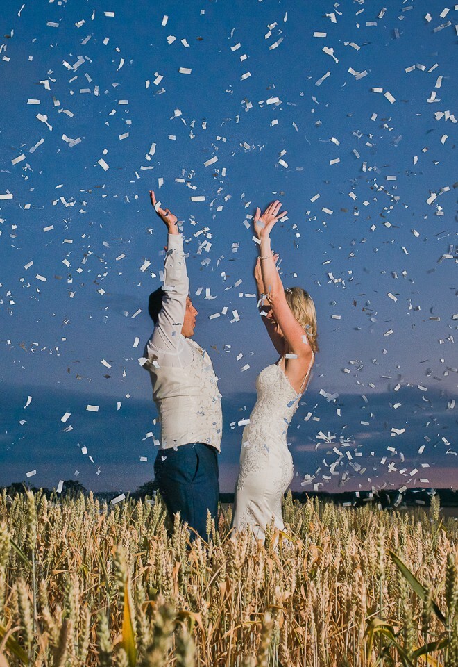 Bride and groom in wheat field during the summer at sunset reach into a mass of silver confetti