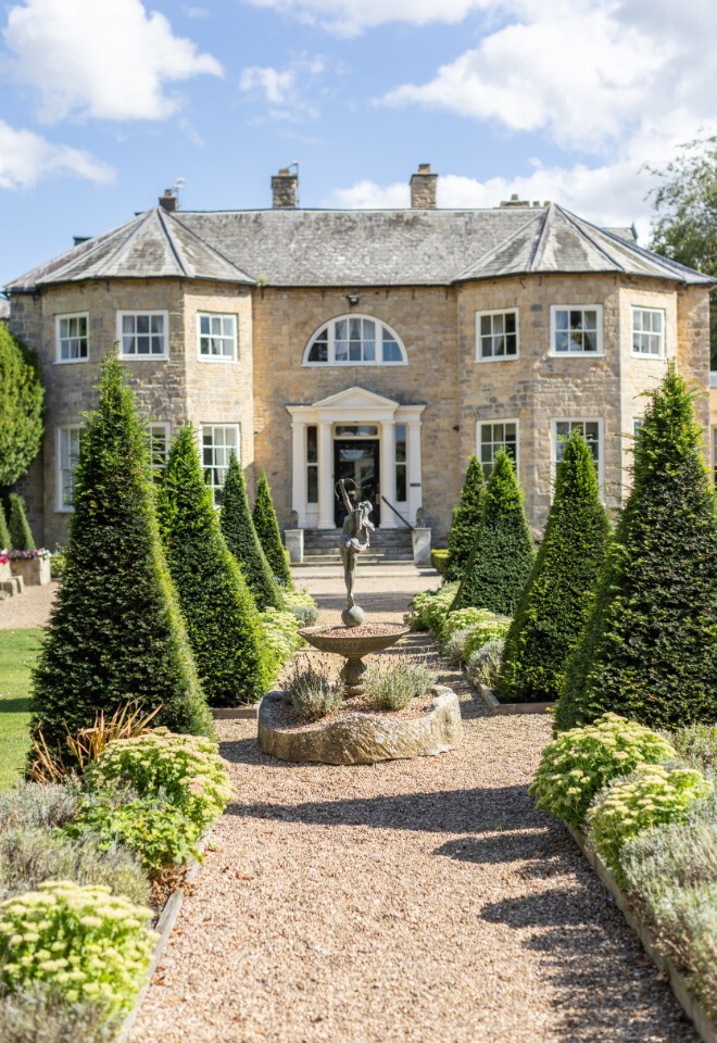 Front view of Washingborough Hall Hotel with a gravel path lined by manicured topiary trees and a central fountain statue under a bright blue sky.