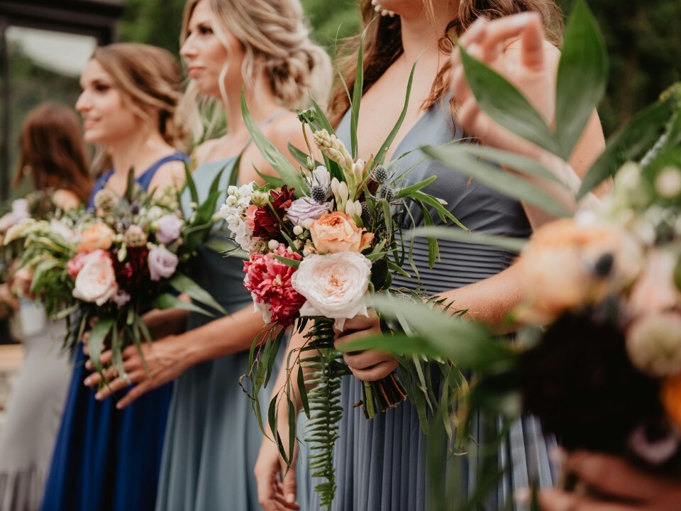 Bridesmaids standing outdoors holding mixed floral bouquets with greenery, wearing blue dresses during a wedding ceremony