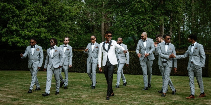 Groom and his entourage (including best man, ushers and groomsmen) walking towards wedding ceremony. They are smiling at each other.