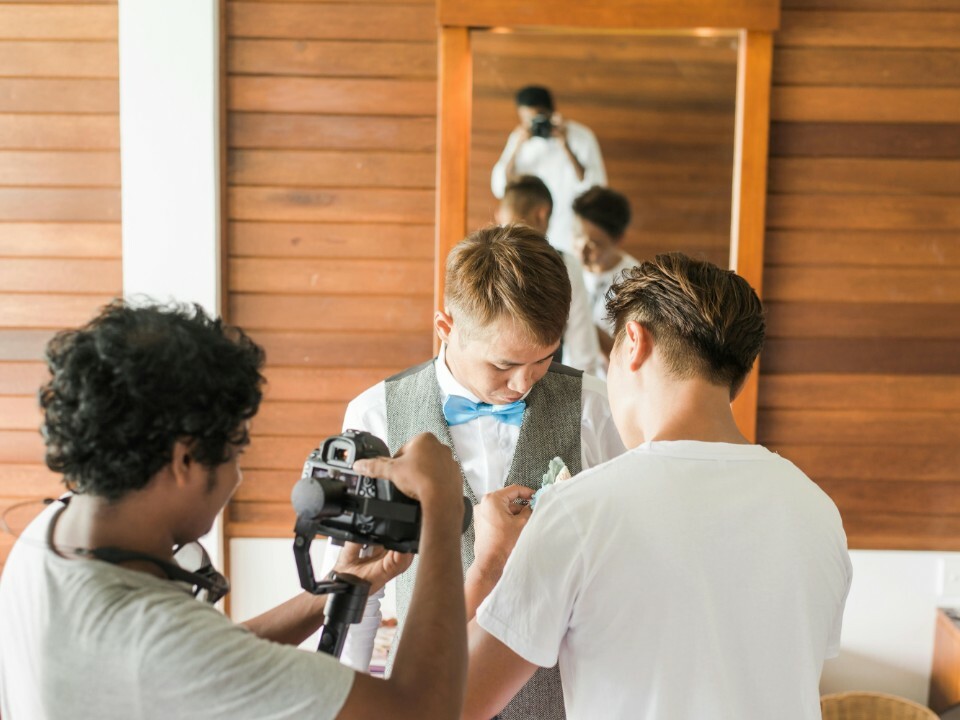 Groom getting ready with help fastening his boutonniere, while a videographer films the moment in a bright, wood-panelled room.
