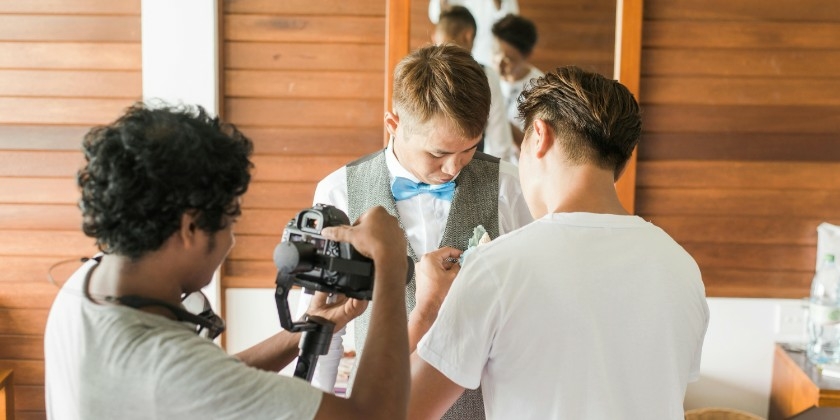 Groom getting ready with help fastening his boutonniere, while a videographer films the moment in a bright, wood-panelled room.