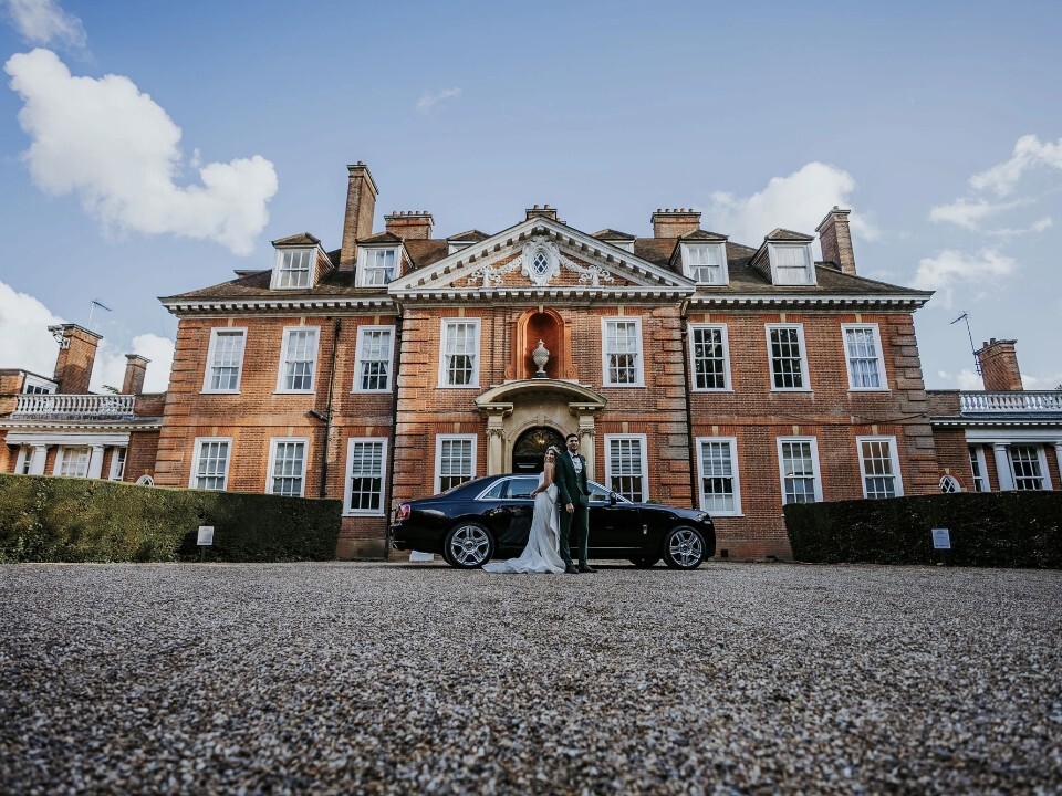 Bride and groom standing in front of Hunton Park Hotel with a luxury car, set against the grand red-brick country house and blue sky