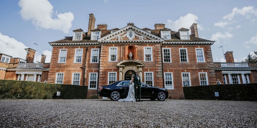 Bride and groom standing in front of Hunton Park Hotel with a luxury car, set against the grand red-brick country house and blue sky