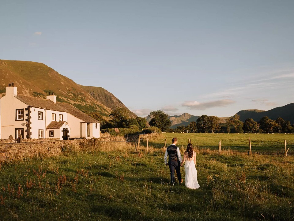 Bride and groom walking hand in hand through fields at New House Farm, with farmhouse and mountain views in warm evening light.