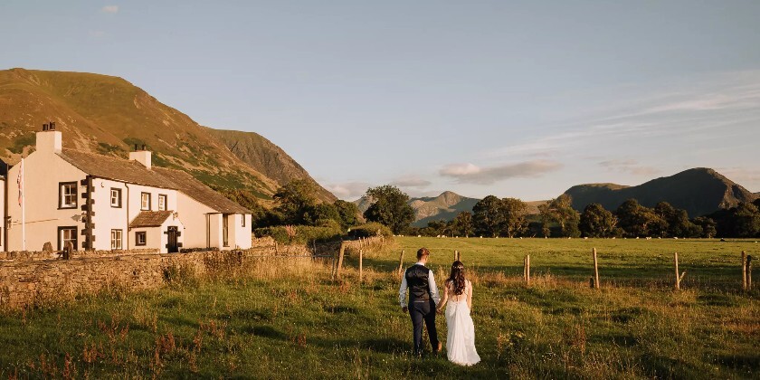 Bride and groom walking hand in hand through fields at New House Farm, with farmhouse and mountain views in warm evening light.