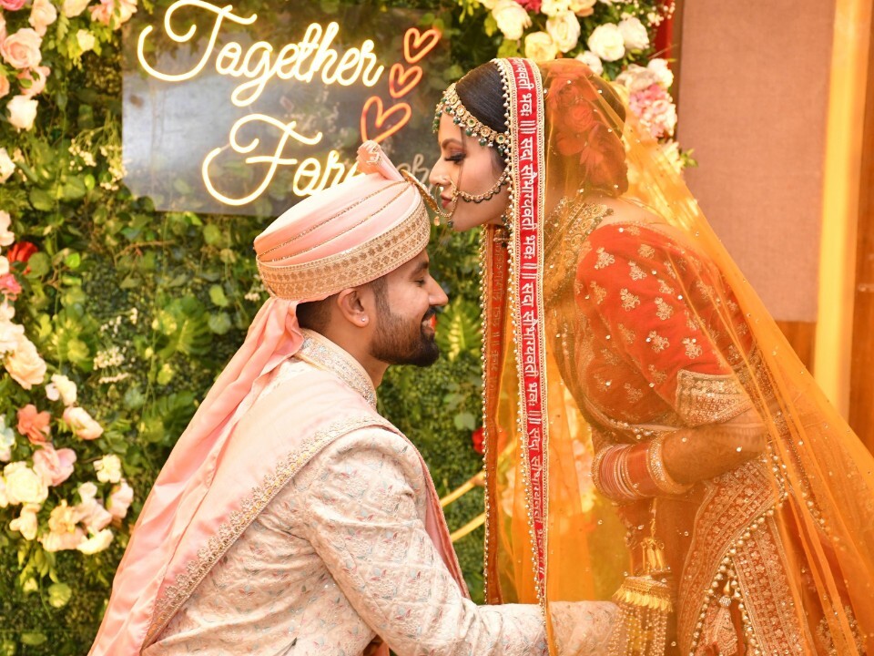 South Asian bride and groom during a wedding ceremony, wearing traditional attire in front of a floral backdrop with a glowing “Together Forever” sign.