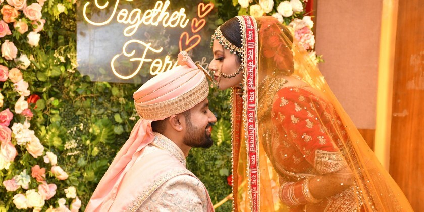 South Asian bride and groom during a wedding ceremony, wearing traditional attire in front of a floral backdrop with a glowing “Together Forever” sign.