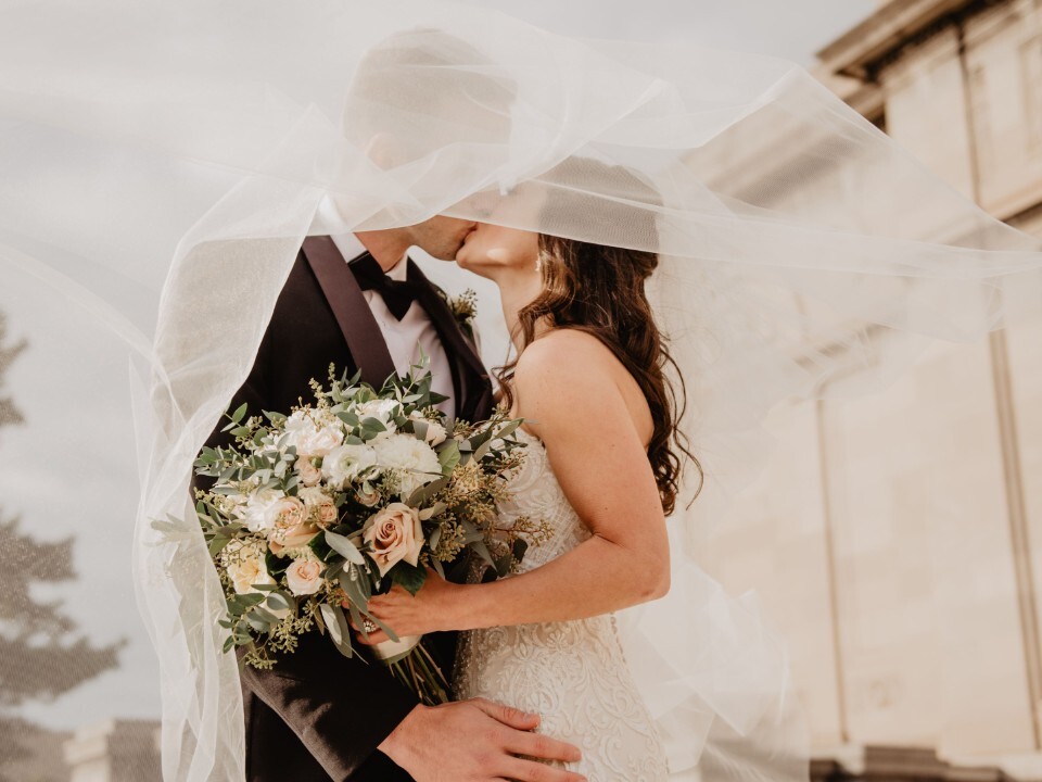 Newly married couple sharing a kiss beneath the brides veil outside wedding venue