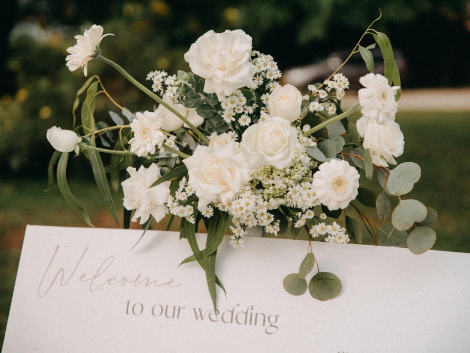 White wedding welcome sign decorated with white roses, daisies and eucalyptus flowers in an outdoor garden setting.