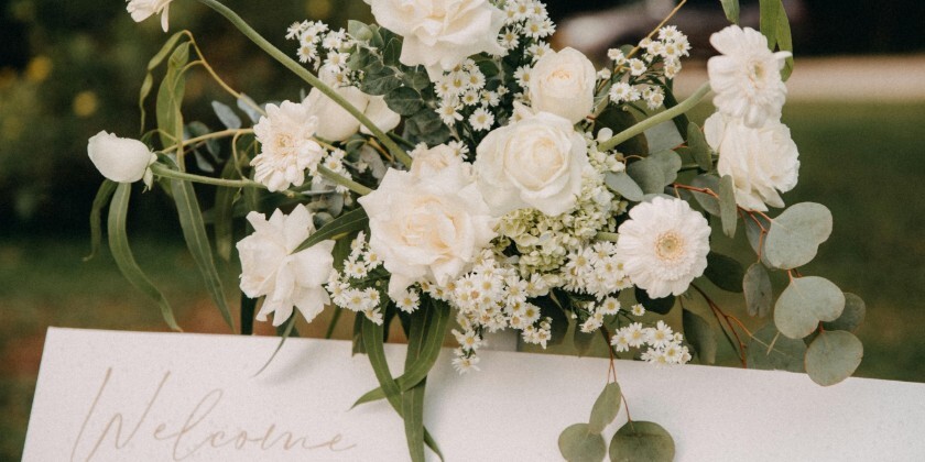 White wedding welcome sign decorated with white roses, daisies and eucalyptus flowers in an outdoor garden setting.