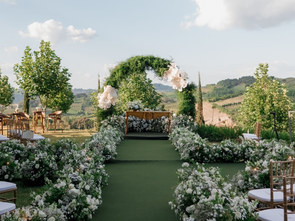 Outdoor luxury wedding ceremony setup with a floral-lined aisle, green arch and panoramic countryside views.
