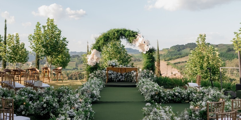 Outdoor luxury wedding ceremony setup with a floral-lined aisle, green arch and panoramic countryside views.