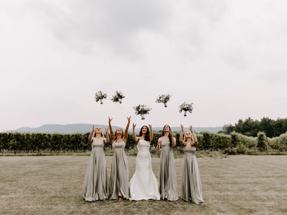 Bride and five bridesmaids in long sage dresses on a lawn throwing white floral bouquets into the air, with countryside and cloudy sky in the background.