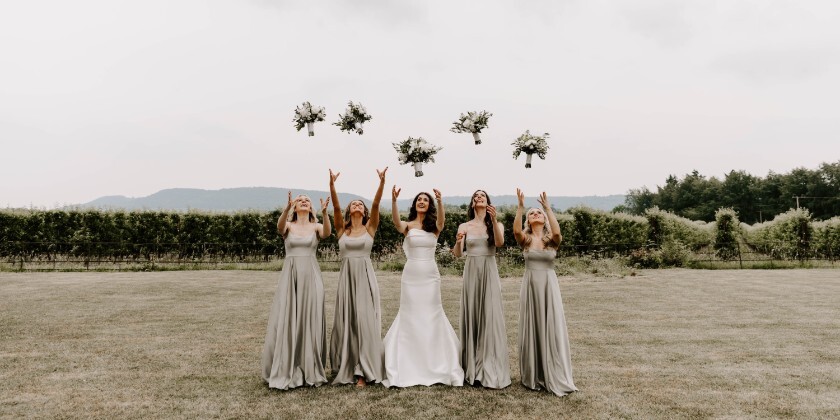 Bride and five bridesmaids in long sage dresses on a lawn throwing white floral bouquets into the air, with countryside and cloudy sky in the background.