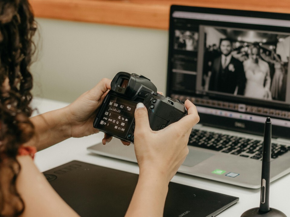 A wedding photographer editing images on a laptop while holding their camera.