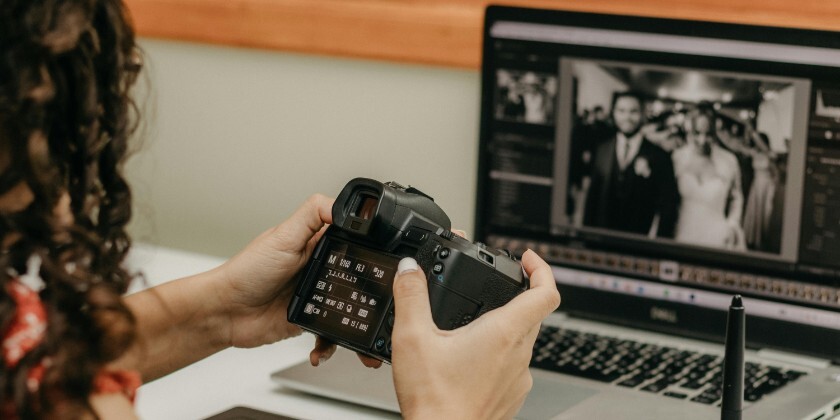 A wedding photographer editing images on a laptop while holding their camera.