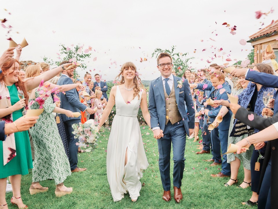 Bride and groom walking through guests throwing confetti at an outdoor wedding, with countryside views and relaxed garden setting.