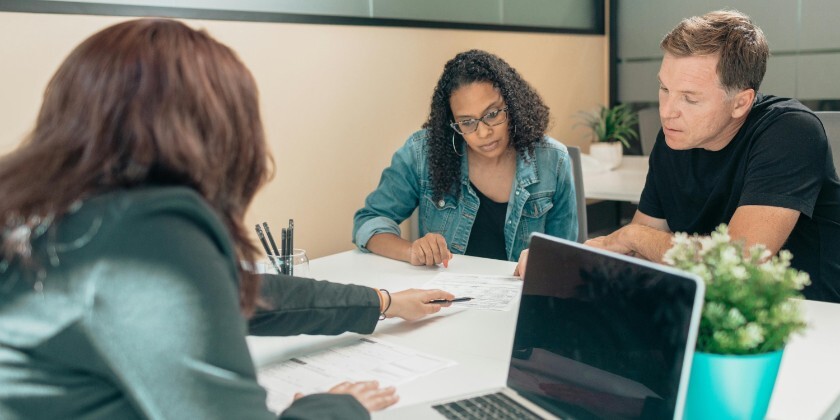 Engaged couple seated at a desk discussing details with another person using a laptop