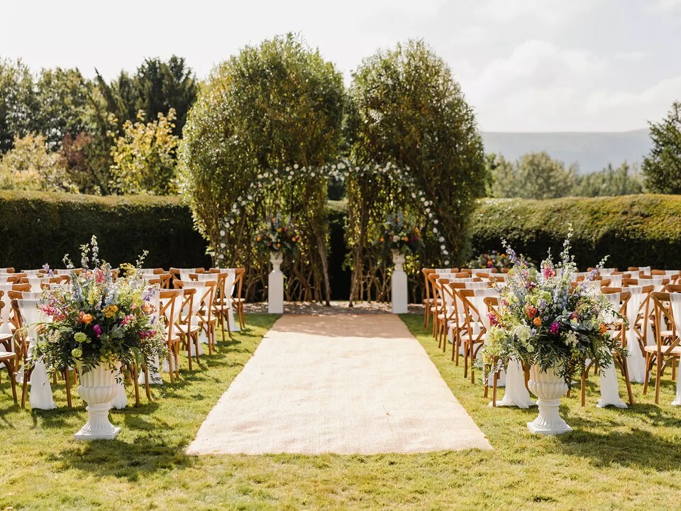 Outdoor wedding ceremony setup at Peterstone Court with floral aisle arrangements, wooden chairs, lawn setting and views of the countryside.