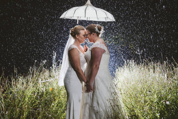 Wedding Photograph of two brides under an umbrella in the rain, raindrops all visible in dramatic lighting