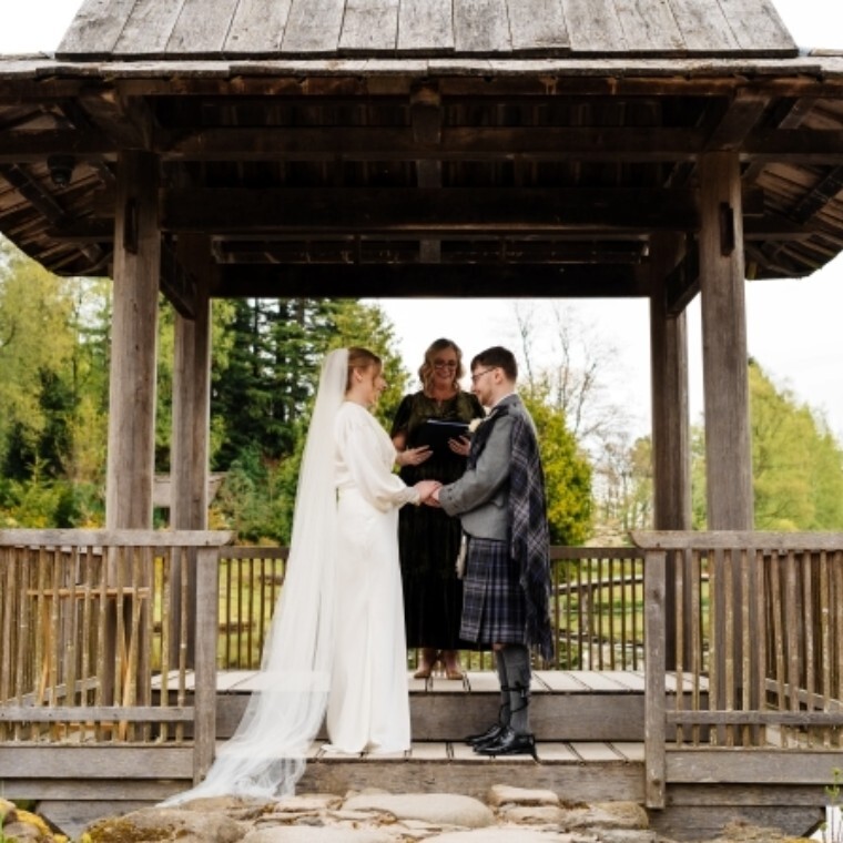 A bride and groom hold hands, gazing at one another with their celebrant behind smiling. They stand on an azumaya in a japanese garden