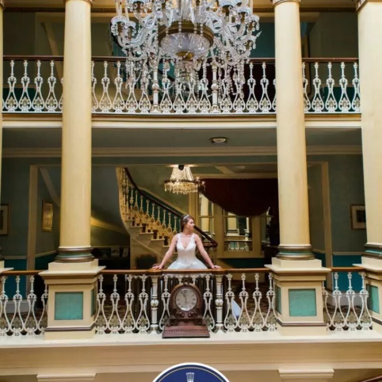 Bride standing by railing with chandelier above and pillars to the side in historic building.
