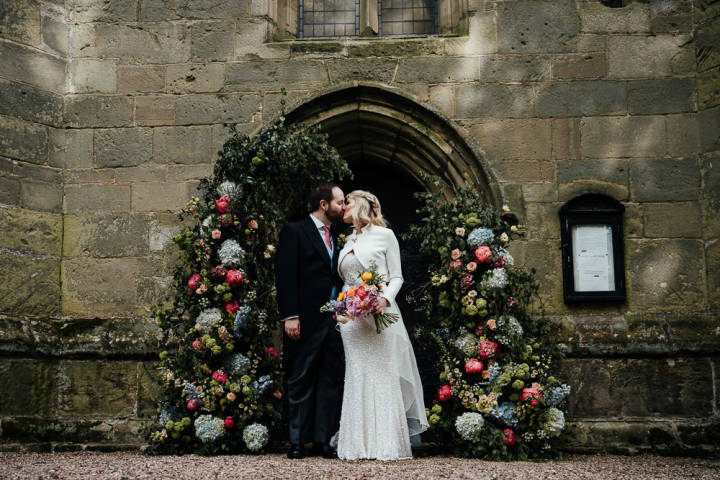 Couple standing and kissing under flower arch outside church 