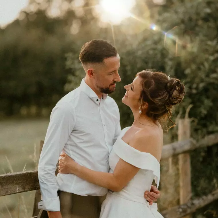 a newly married couple hugging and leaning against a fence at sunset