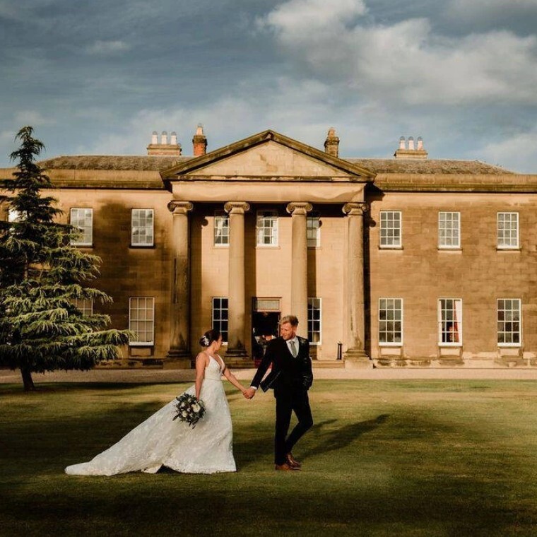 Wedding couple walking through the grounds at Rise Hall wedding venue with the venue in the background.