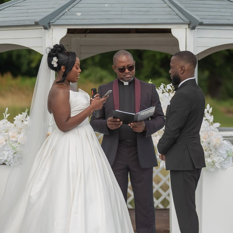 An outdoor wedding ceremony with a bride and groom standing beside a pastor under a decorated white gazebo. White flowers and greenery surround the ceremony area.