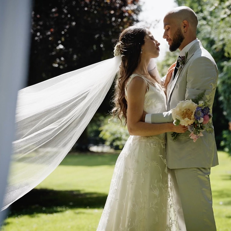 A bride in a lace gown and long veil embraces her groom in a light grey suit during a sunlit outdoor garden wedding, framed in the foreground by their white aerial silks.