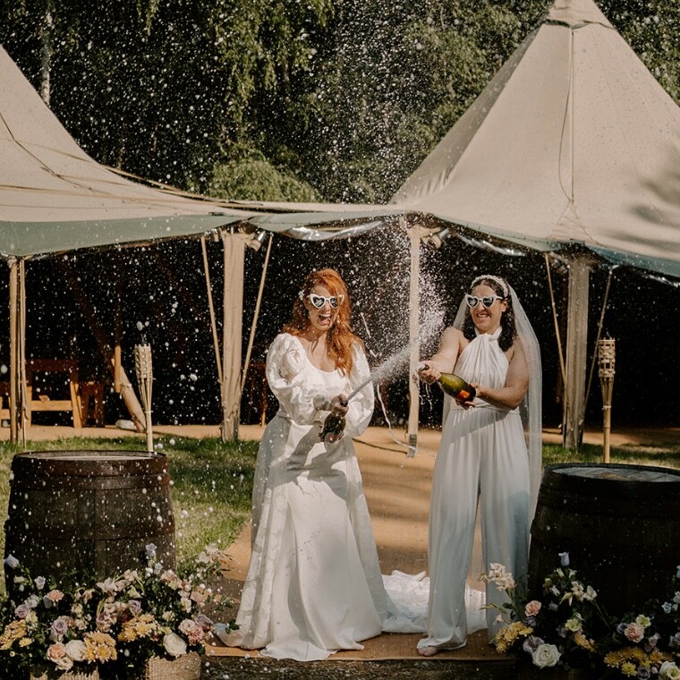 Two brides popping champagne infront of a 2 hat tipi
