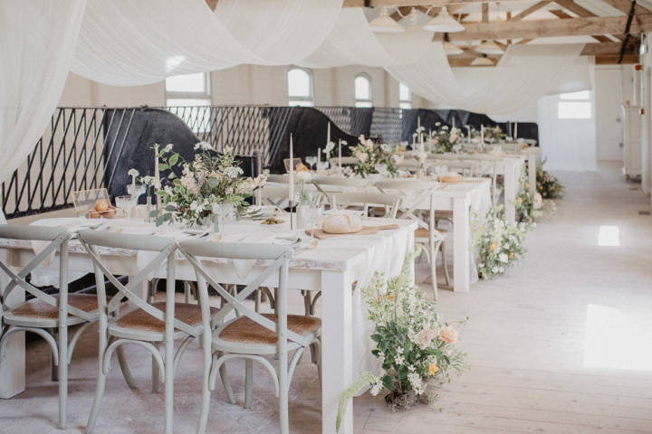 Renovated barn dining set up for a wedding with white drapes hanging over the beams and pastel flowers