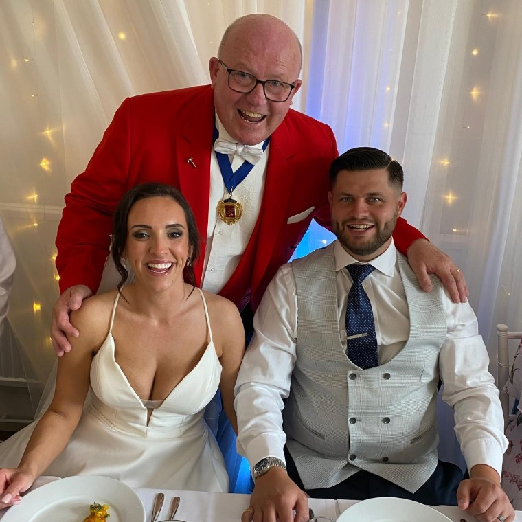 Surrey wedding toastmaster Toastmaster John in his distinctive red tailcoat posing with a smiling bride and groom during their wedding breakfast.