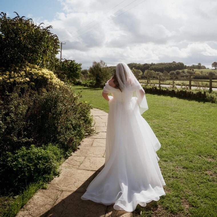 a bride walking along a paved footpath surrounded by fields with her vale and train flowing behind her