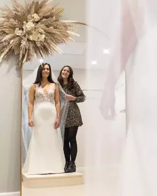 Bride in wedding dress at bridal boutique looking in mirror with consultant. Dress has lace and beading with a plunge vneck.