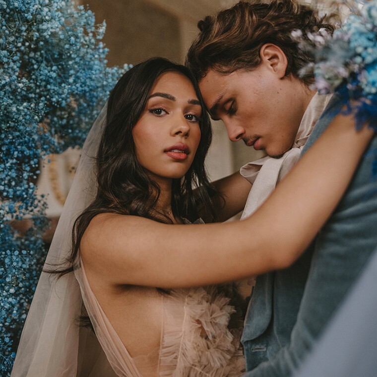A couple embrace on their wedding day, the bride, wearing a ruffed tulle dress looks into the lens of the camera whilst her arms are draped round the neck of her groom dressed in a blue velvet jacket.