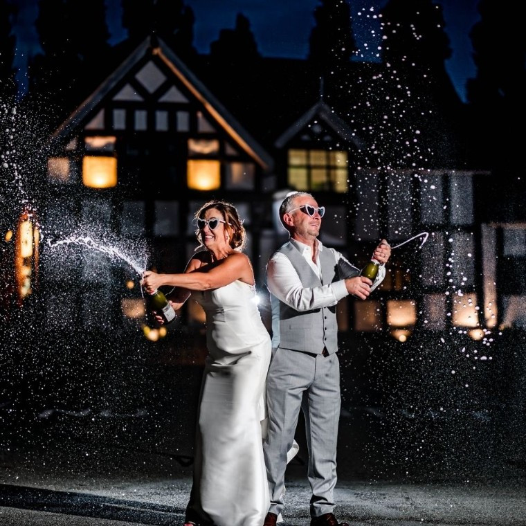 Newlyweds celebrating with a champagne spray outside The Cock OBarton. an award-winning countryside wedding venue in Cheshire.