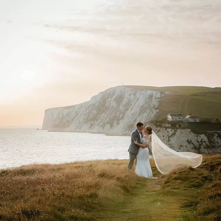 bride with veil flowing in the wind being held by her new husband facing each other at dusk on a cliff top over looking a bay